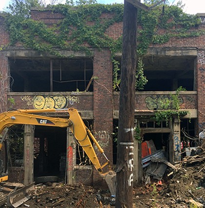 excavator in front of abandoned building with ivy on it and telephone pole in foreground