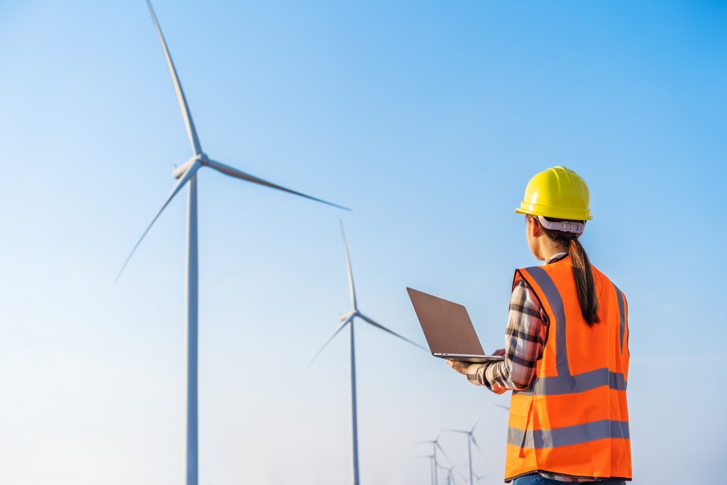 female worker in helmet and vest looking on at wind turbines against a blue sky