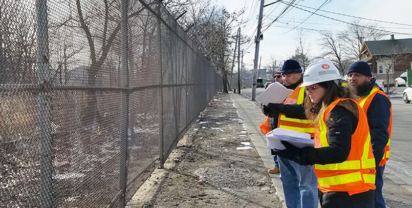 people in high vis vests and hard hats taking notes on the street next to a chain link fence and trees