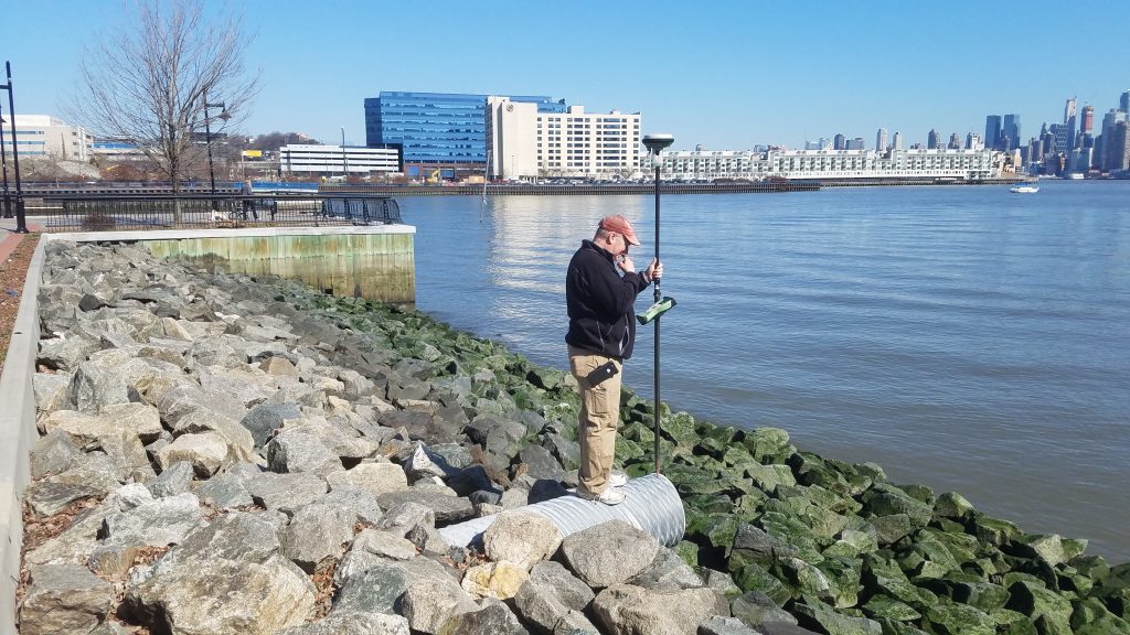 employee using survey equipment on a rocky shore with buildings in the background
