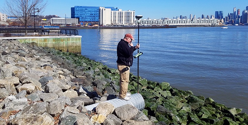 man holding survey equipment on rocky shore by water with buildings in distance