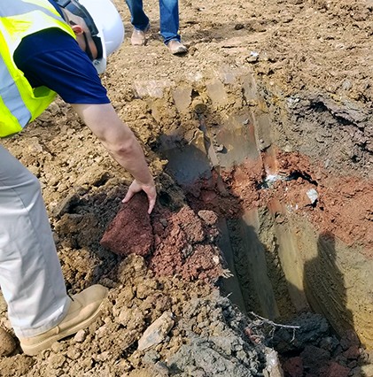 worker bent over by open hole in soil where soil striations are visible, his hand is on large dirt rock