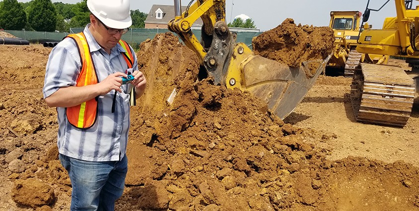 worker in hard hat and vest looking at digital camera in front of large pile of dirt and excavators