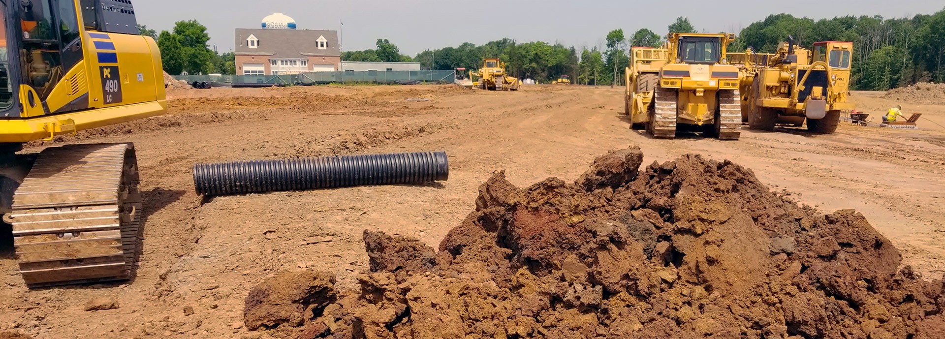 excavators around pile of dirt and pipe with buildings in distance
