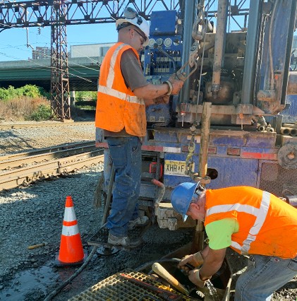 2 workers in vests and hard hats working with drill equipment on train tracks