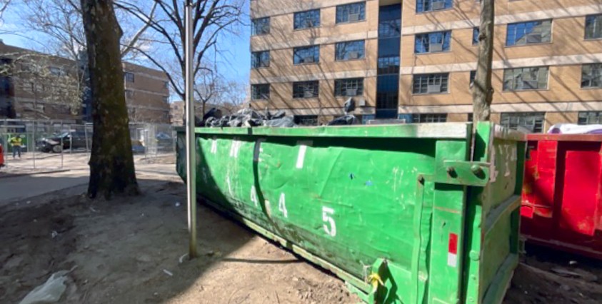 green dumpster on dirt near tree and building in background