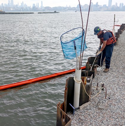 worker by water at marina with blue net and boom in water and skyline in distance