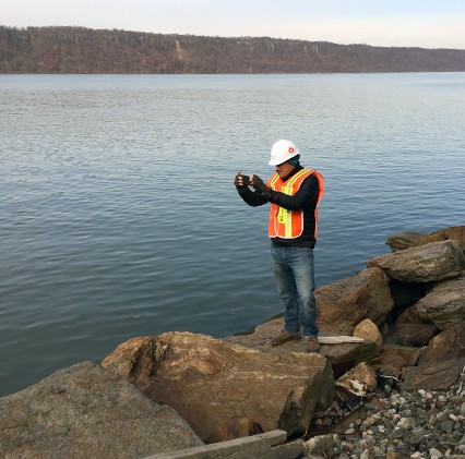 worker in hard hat and vest on rocky shore taking photo in front of river