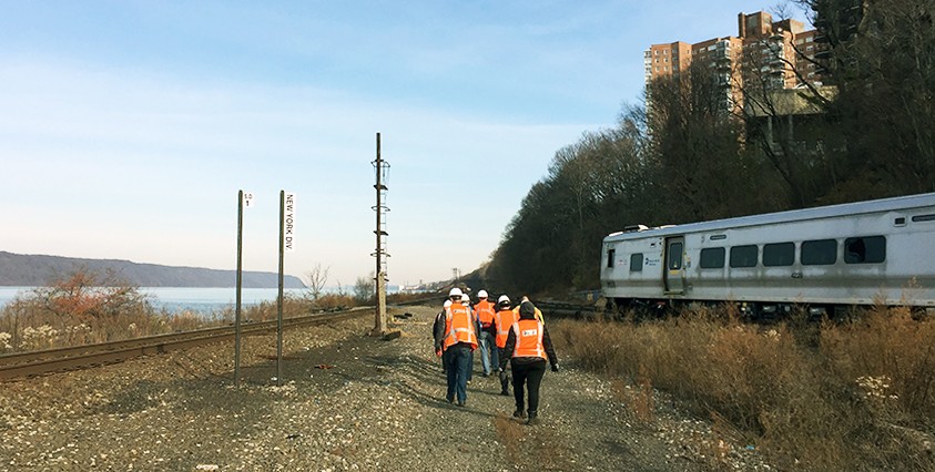 workers in vests walking along train tracks with river on left and buildings and train on right