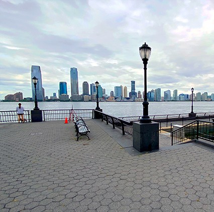 view of boardwalk pier of battery park with lamp post and benches with jersey city in distance