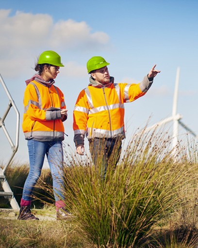 two field workers in hardhats walking on a site with one man pointing and a woman holding a clipboard with a wind turbine in the background