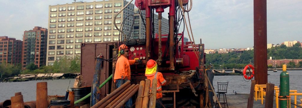 2 workers in the center of a barge with a drill on the hudson with buildings in the background