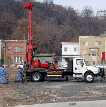 geotech drilling equipment and truck with workers in a dirt field with houses in the background